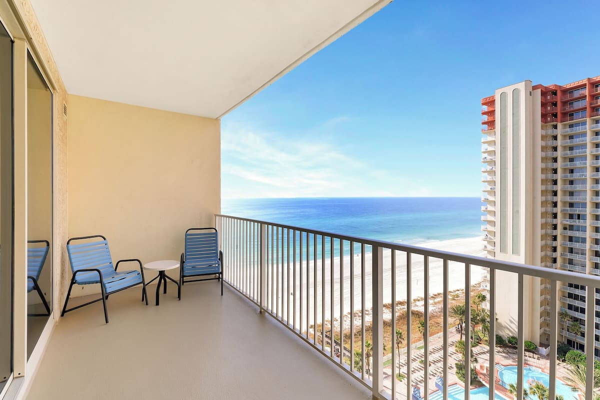 A balcony with two blue chairs and a small table, overlooking a beach and a pool area.