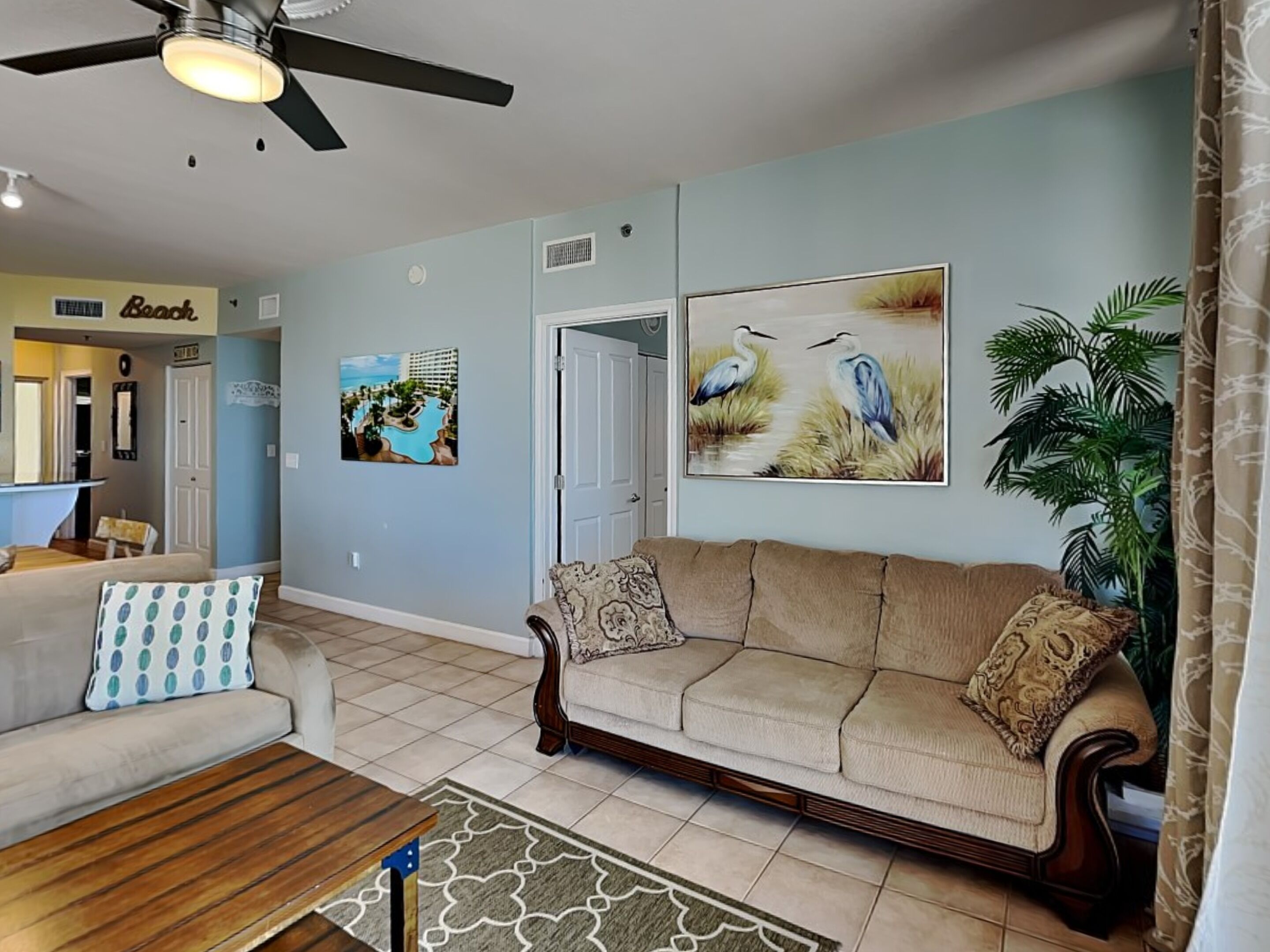 A living room with a beige sofa, a wooden coffee table, and a patterned rug. There are two wall-mounted paintings, one of a beach resort and another of birds. A ceiling fan is visible, and a plant is in the corner.