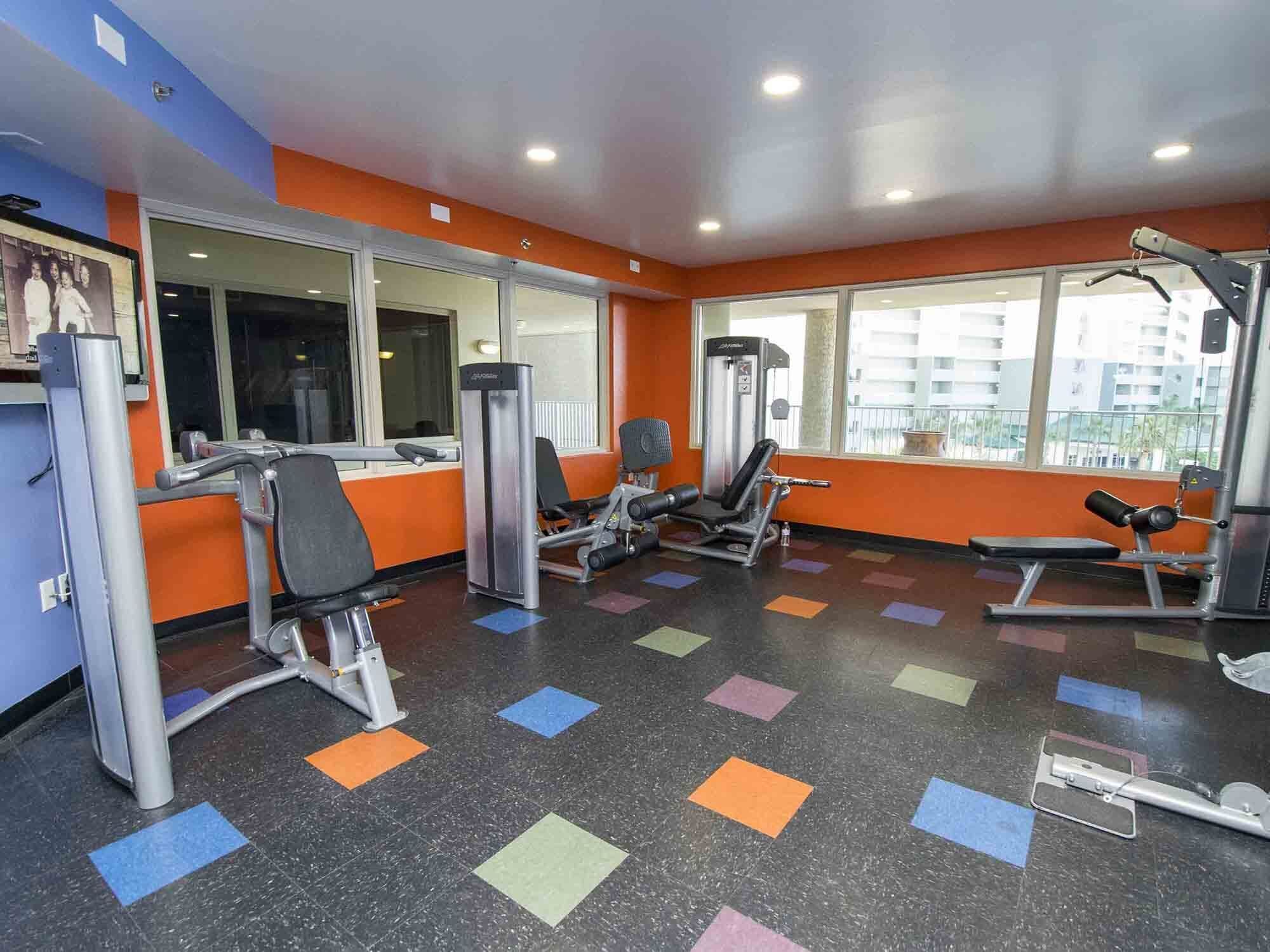 Un gimnasio con diversas máquinas de ejercicio, ventanas amplias y un suelo colorido.
