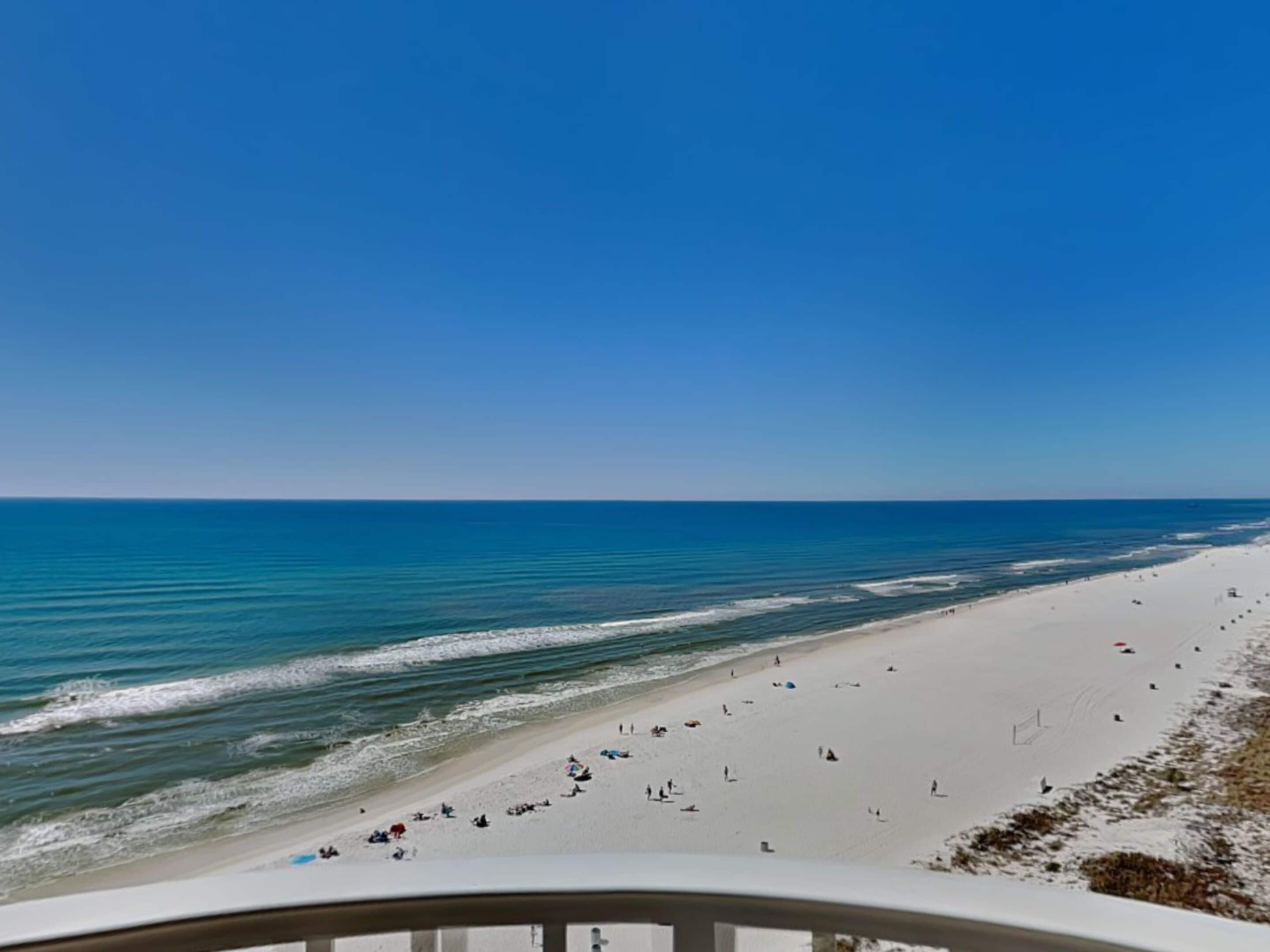 A beach view with a clear sky, calm sea, and a sandy shore with people and umbrellas.