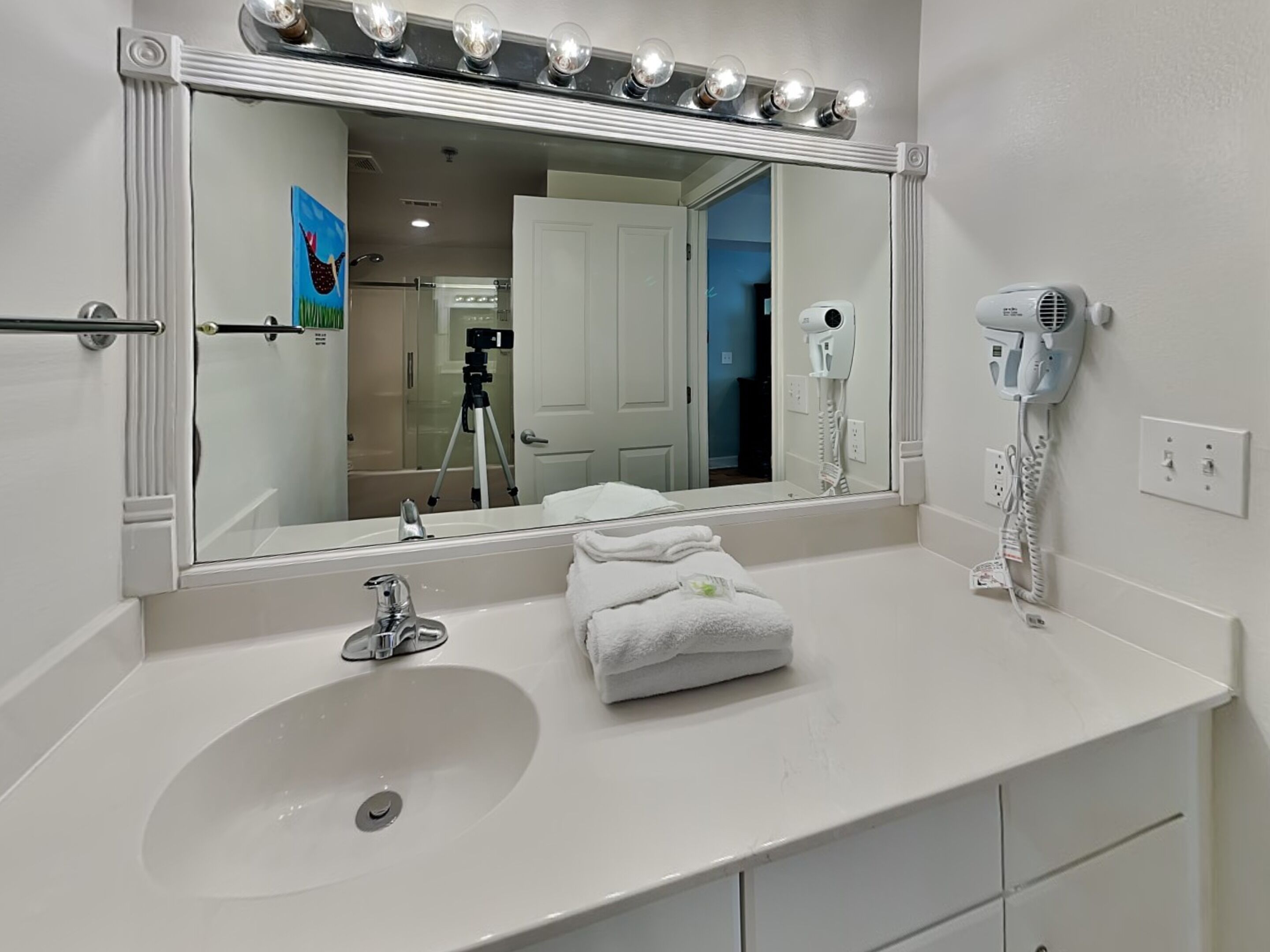 A bathroom with a large mirror, vanity with a sink, a hairdryer, and a tripod in the background.