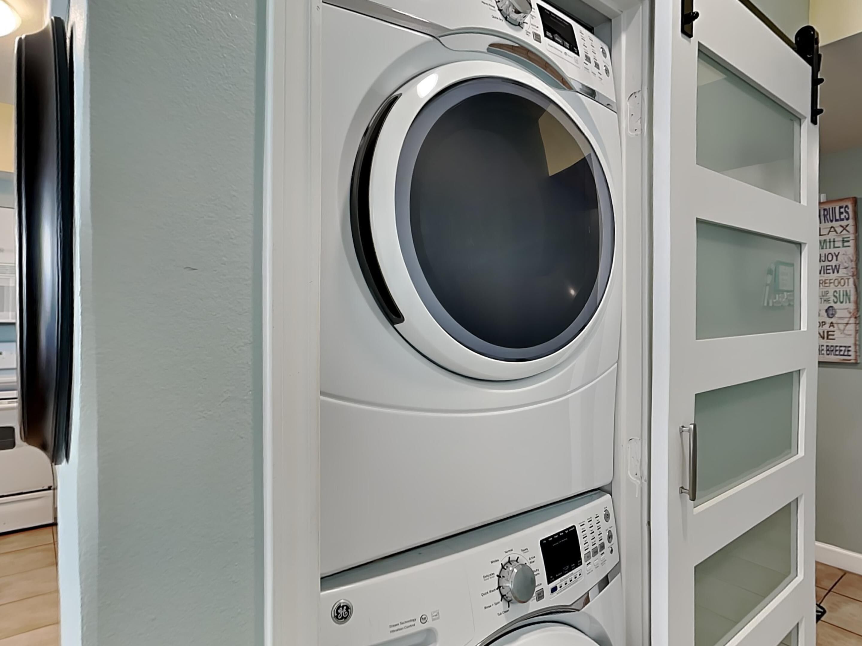 A front-loading washing machine with a control panel, a dryer, and a glass-fronted cabinet with a poster.