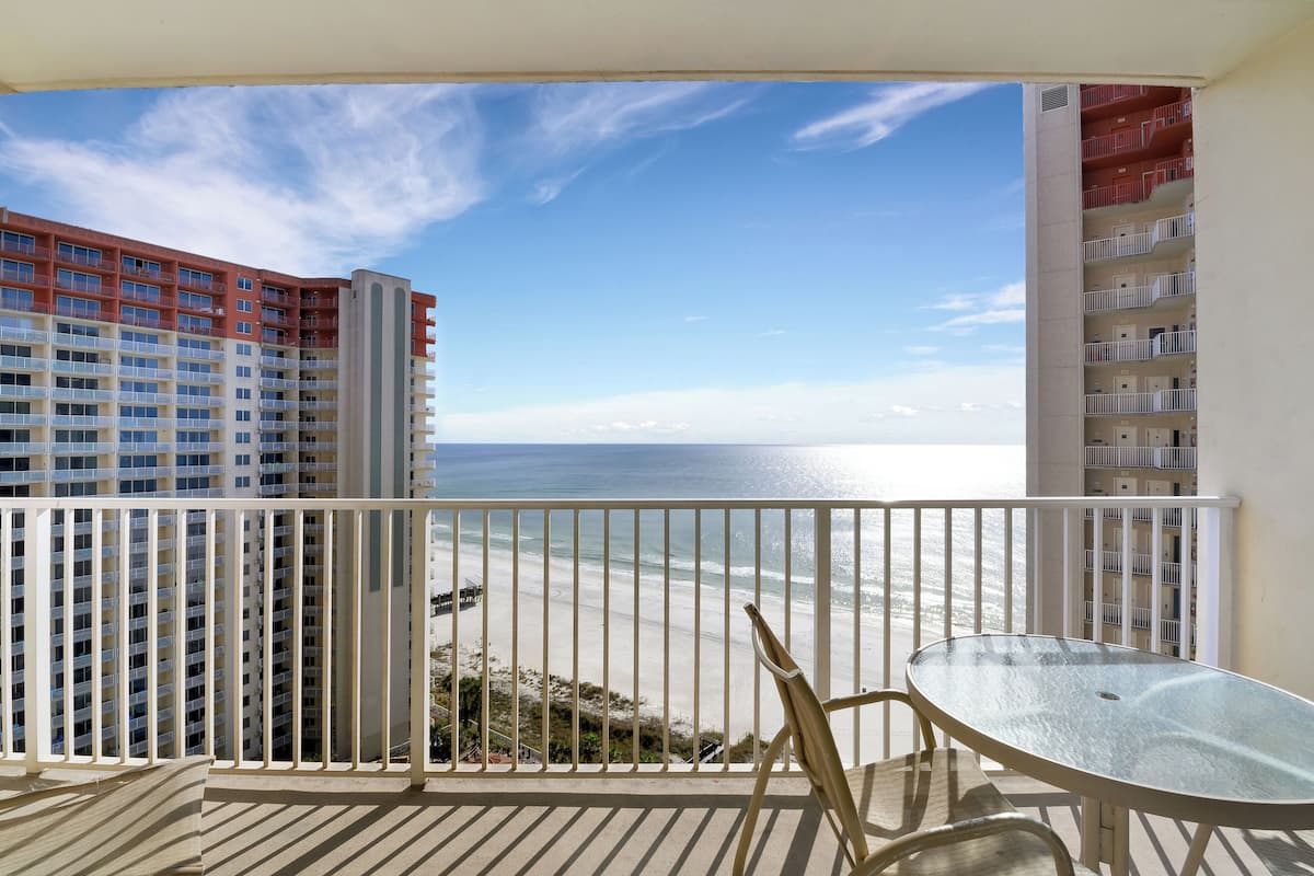 A balcony with a glass table and chairs overlooking a beach and a high-rise building.