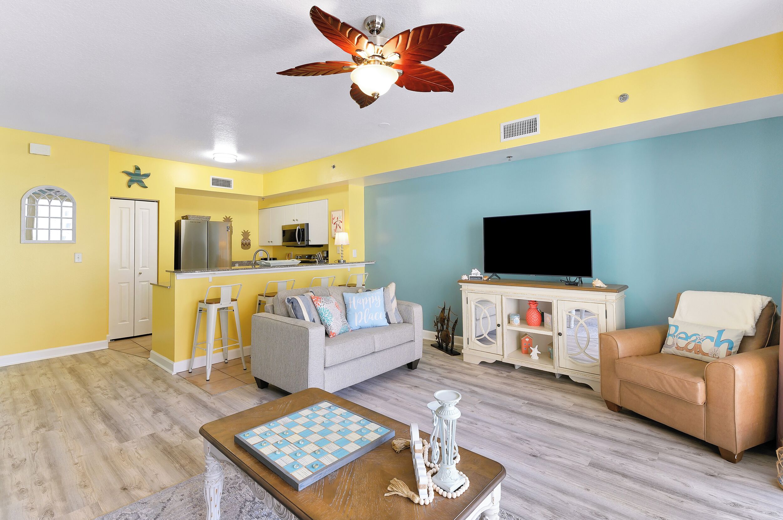 A living room with a sofa, a coffee table, and a television. There is a kitchen area with a refrigerator, microwave, and a dining table. The room has a ceiling fan and a window.