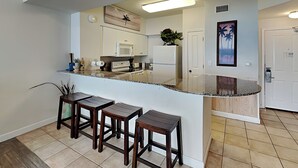 A modern kitchen with a central island, bar stools, a microwave, and a refrigerator. - Shores of Panama III by Resort Collection (Panama City Beach)