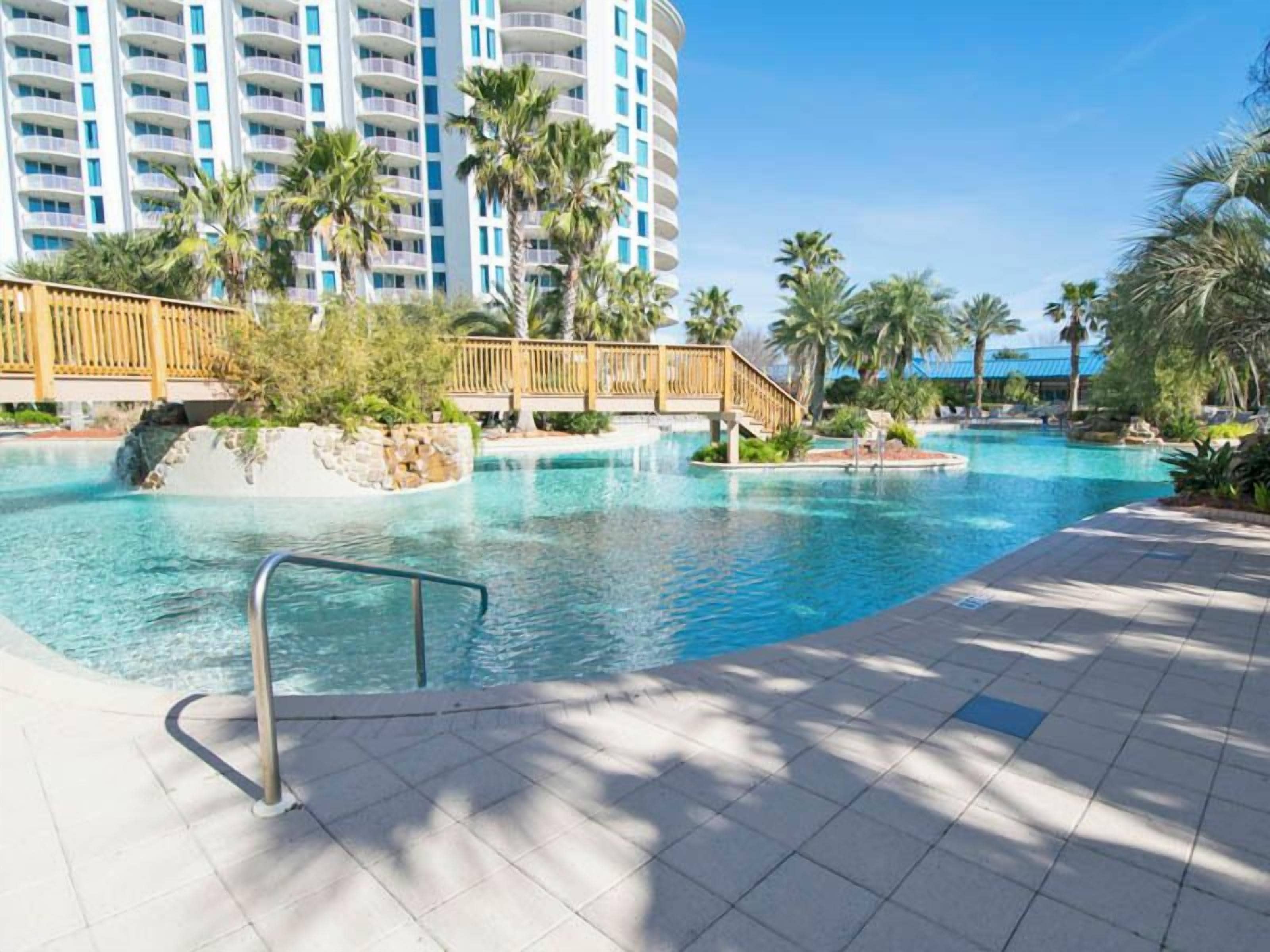 Zona de piscina en un resort con un puente de madera, palmeras y un edificio alto al fondo.