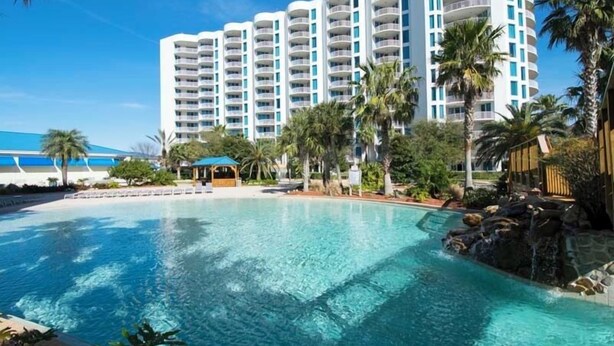 A large swimming pool with clear blue water, surrounded by palm trees and a multi-story hotel building with balconies. - The Palms of Destin by Southern Vacation Rentals (Destin)
