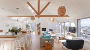Een ruime woonkamer met een groot raam, een houten plafond en een moderne keuken met witte barstoelen.