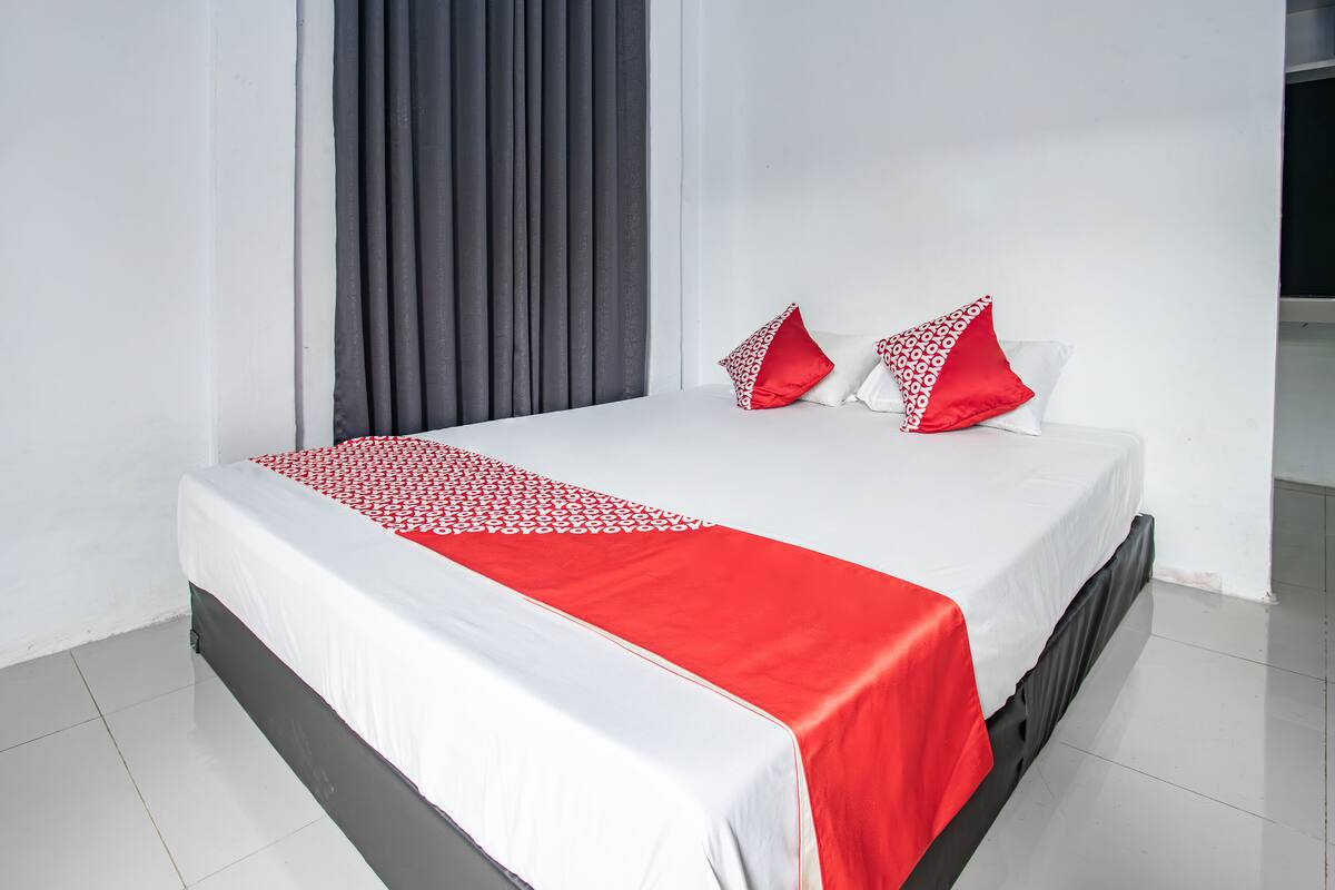 A neatly made bed with a white and red bedspread, two pillows, and a black headboard in a room with white walls and a grey curtain.