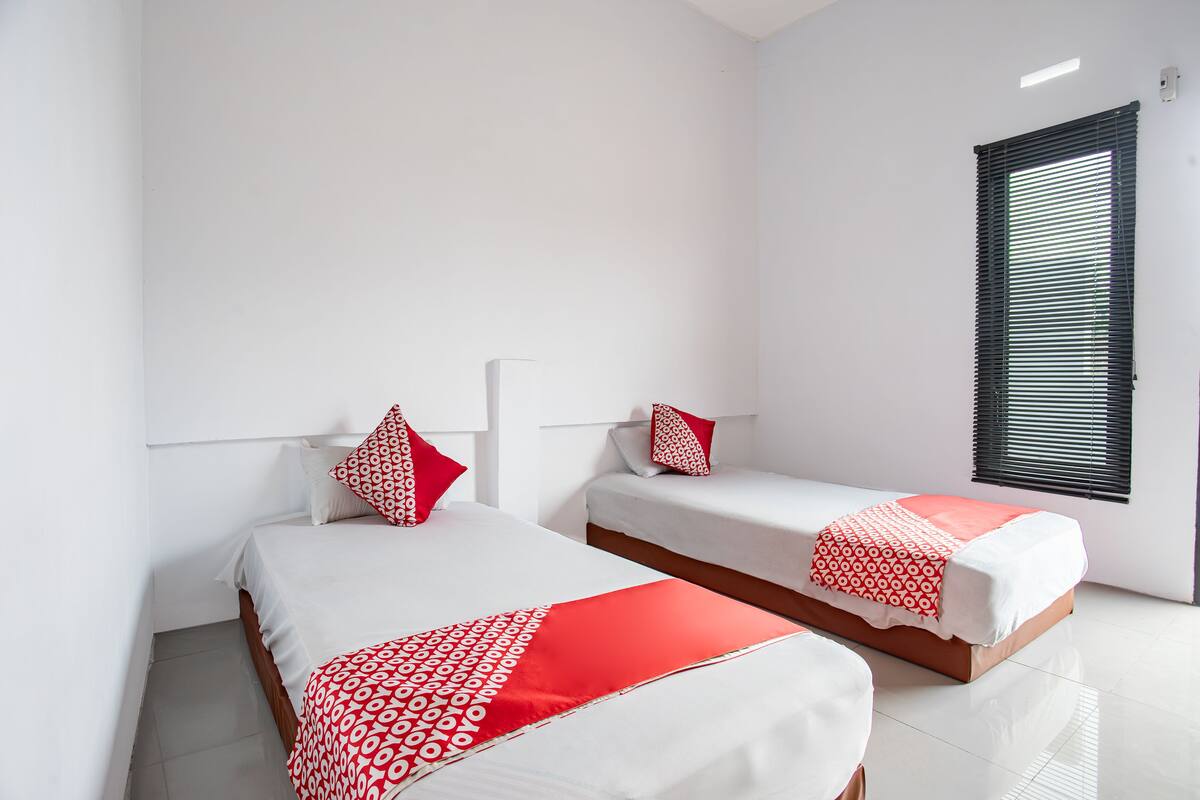 A hotel room with two beds, each with a white and red patterned bedspread, and a window with blinds.