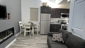 Une cuisine moderne avec des meubles de couleur sombre, un réfrigérateur en acier inoxydable et un four à micro-ondes. On y trouve un coin repas avec une table et des chaises blanches, ainsi qu’un canapé gris agrémenté d’un coussin à motifs.