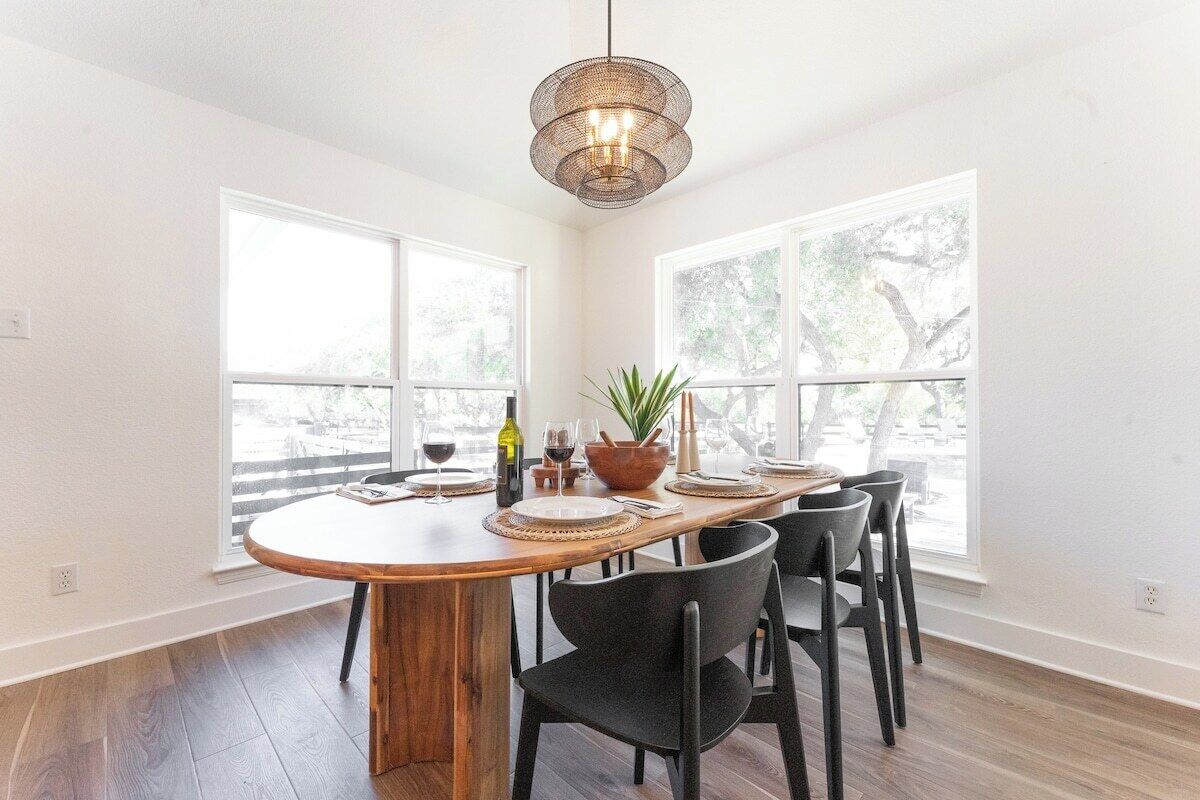 A dining area with a wooden table, black chairs, a woven pendant light, and large windows with a view of trees.