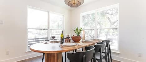 Un espace repas comprenant une table en bois, des chaises noires, un luminaire suspendu en fibres tressées et de grandes fenêtres donnant sur la forêt.