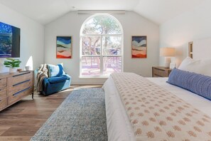 A bedroom with a large window, a bed with patterned bedding, a blue chair, a wooden dresser, and artwork on the walls. - Secluded Hill Country Luxe: Vibes, Pool, Best Spot (Austin)