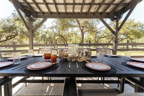 An outdoor dining area with a table set for two, surrounded by a wooden fence, and trees in the background. - Secluded Hill Country Luxe: Vibes, Pool, Best Spot (Austin)