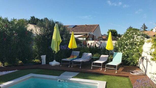 Piscina com deck de madeira, cercada por vegetação e uma casa com telhado de telhas.