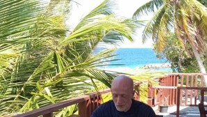 Un homme est assis à une table en plein air, devant une boisson, une assiette de nourriture et un arrangement floral.