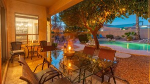 An outdoor dining area with wicker chairs and a glass table, overlooking a swimming pool and a tree with hanging lights. - Luxury 5 Bedroom Scottsdale Home with Heated Pool! (Scottsdale)