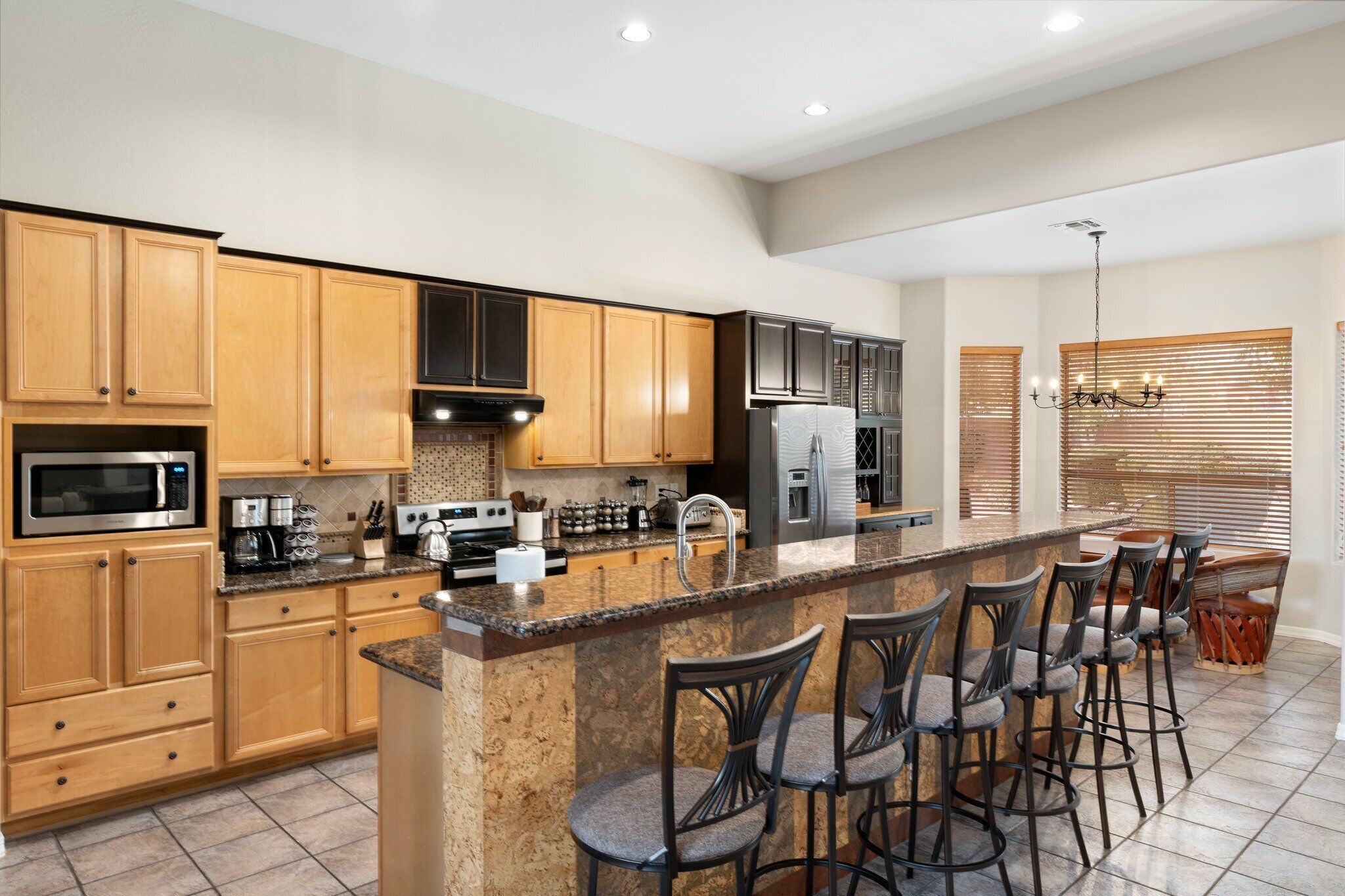 A modern kitchen with wooden cabinets, granite countertops, a central island with bar stools, stainless steel appliances, and a chandelier.