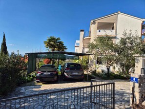 A residential building with two cars parked in front, a covered parking area, and a stone pathway leading to the entrance. - Holiday flat with sea view (Njivice)