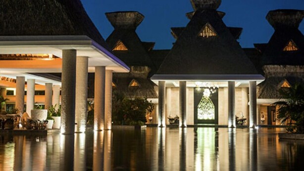 A hotel with a modern design, featuring a central atrium with a reflective floor, large pillars, and a thatched-roof entrance.