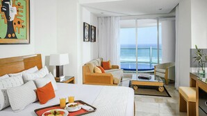 A hotel room with a large bed, a sofa, a coffee table, and a dining table with breakfast. There is a large window with a view of the ocean.
