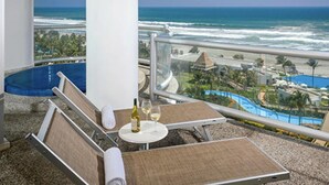 A balcony with a view of the ocean, a pool, and a lounge area with a table and chairs.