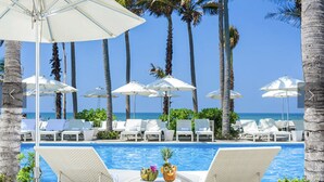 A poolside area with lounge chairs and umbrellas, palm trees, and a clear view of the ocean.
