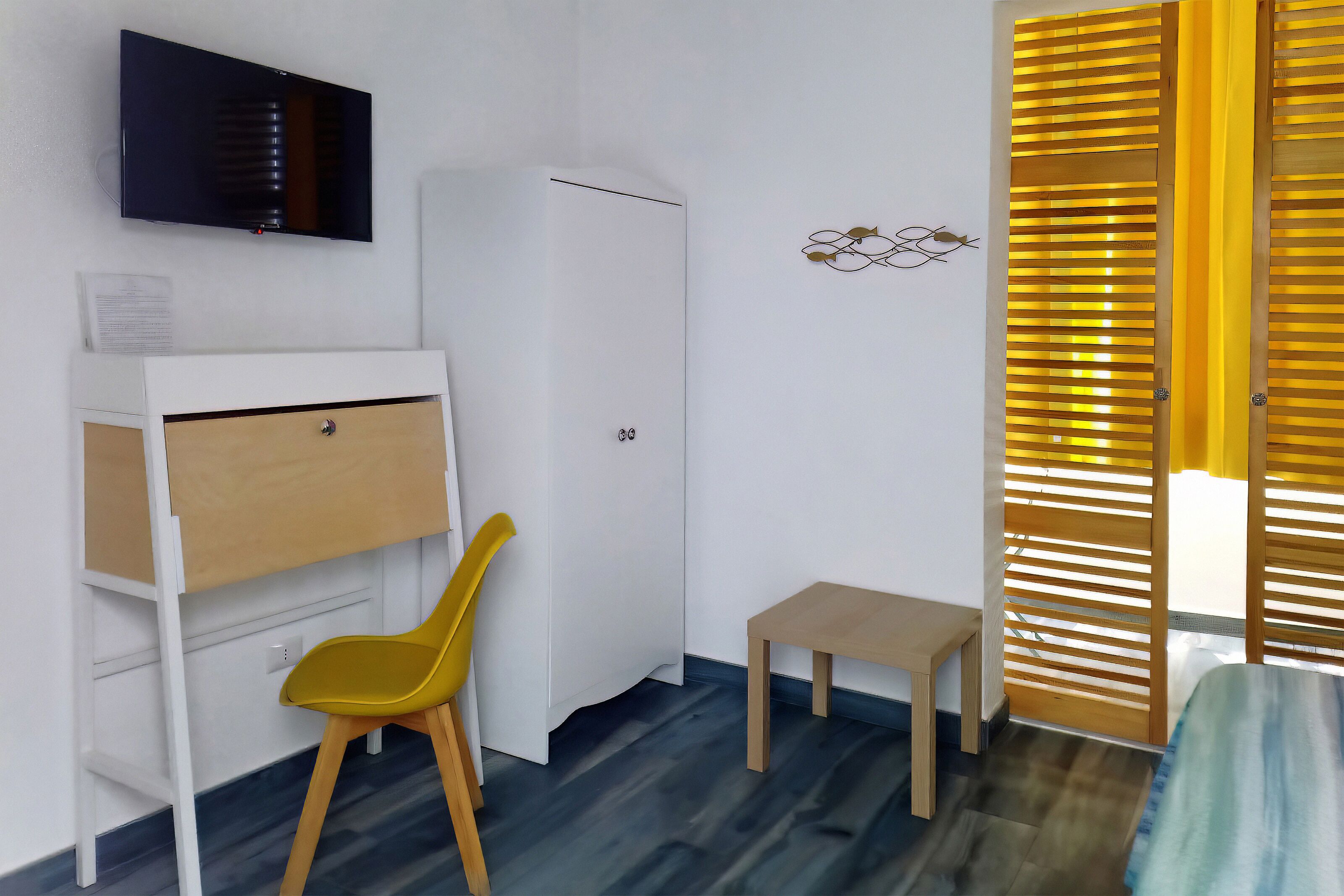 A room with a white wardrobe, a small wooden table, a yellow chair, and a TV mounted on the wall.