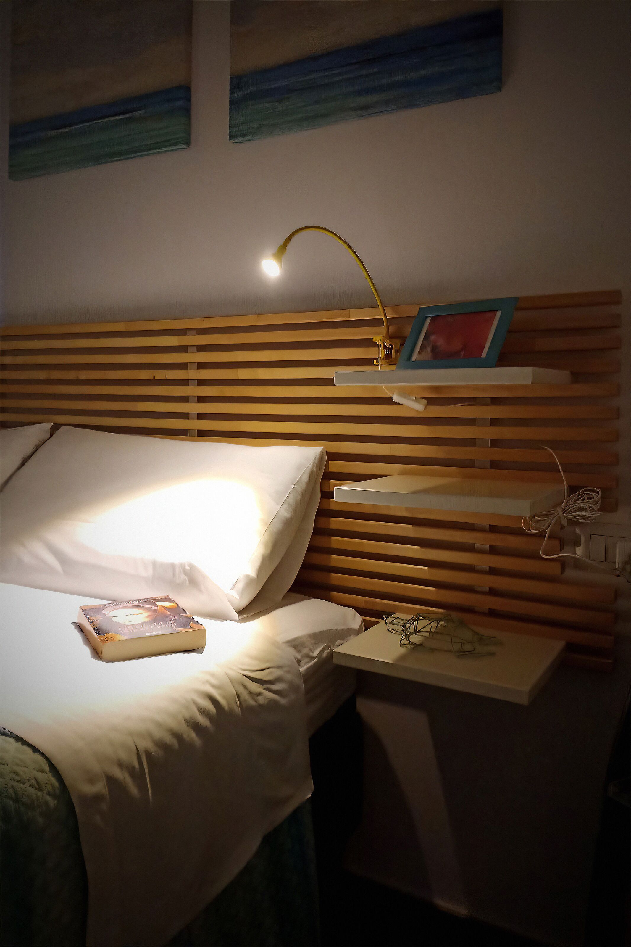 Una cama bien tendida con un almohadón, una mesita de noche con una lámpara y una pequeña estantería con un marco de fotos.
