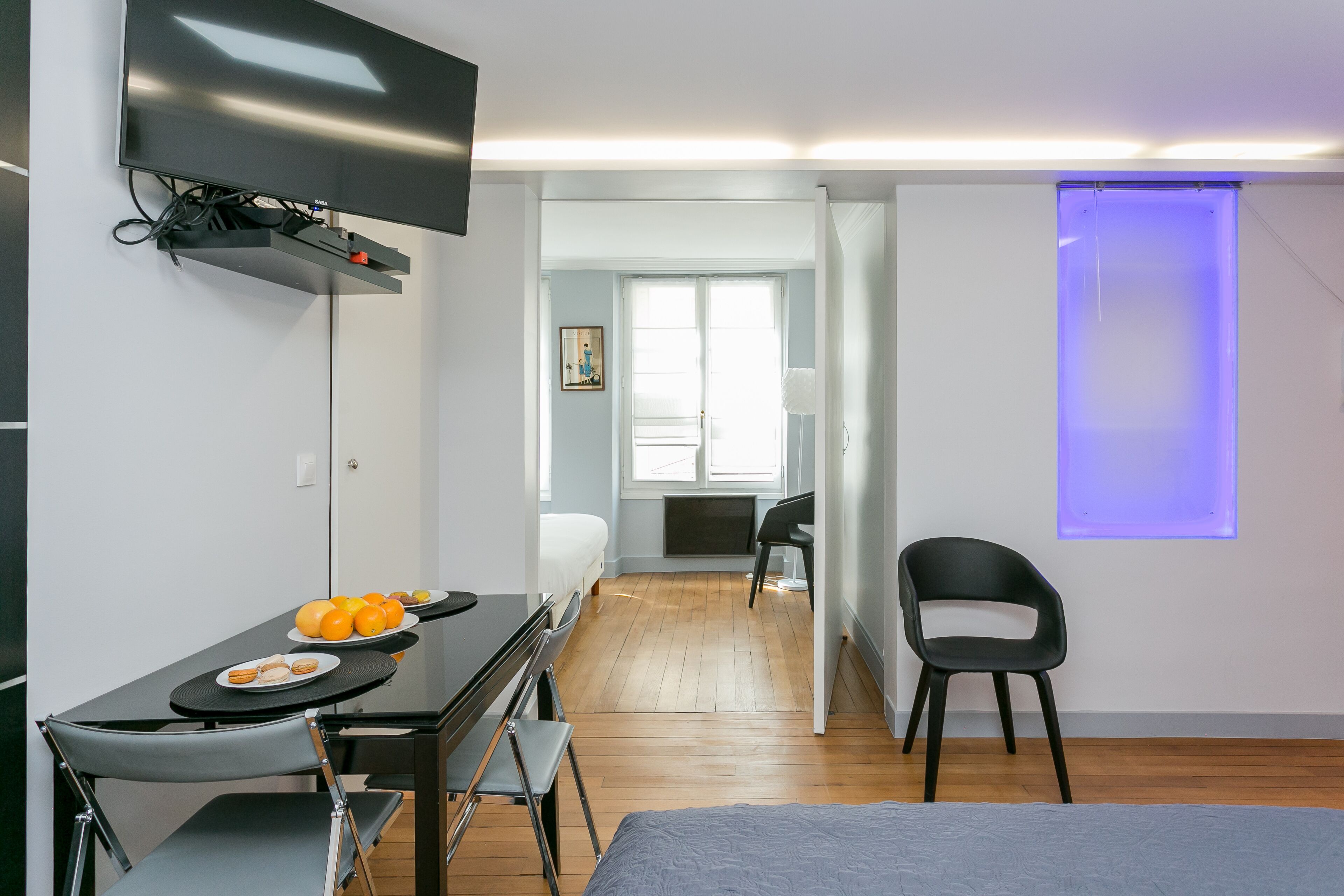 A modern room with a bed, a desk with a chair, a flat-screen TV mounted on the wall, and a dining table with chairs.