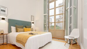 Une chambre moderne avec un grand lit, une table de chevet, une chaise et une vue sur la ville grâce à de grandes fenêtres.