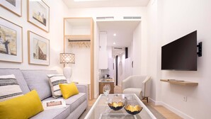 Una sala de estar moderna con un sofá gris, cojines amarillos, una mesa de centro de cristal y un televisor de pantalla plana montado en la pared.