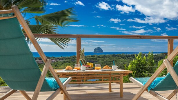 A wooden deck with a table set for breakfast, overlooking a scenic view of the ocean and a distant island. - Manguier (Le Diamant)