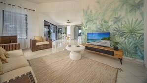 A modern living room with a large TV, a sofa, a coffee table, and a wall mural of tropical leaves. - Manguier (Le Diamant)