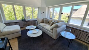 A spacious living room with a large window, a beige sofa, two round tables, and a dark carpet. - Le Bijou Bahnhofstrasse / Paradeplatz LB18B (Zürich)