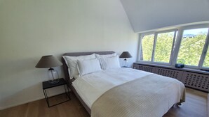 A bedroom with a large bed, two bedside tables with lamps, a window with a view of greenery, and a sloped ceiling. - Le Bijou Bahnhofstrasse / Paradeplatz LB18B (Zürich)