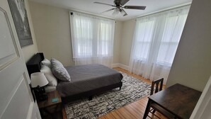 Un dormitorio con cama, escritorio, silla, ventilador de techo y dos ventanas con cortinas translúcidas.