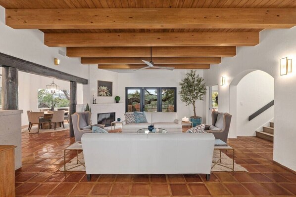 Uma sala de estar ampla com um sofá branco, vigas de madeira no teto, uma lareira e vista para o exterior.