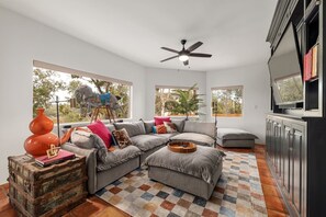 A modern living room with a sectional sofa, a vintage trunk, a ceiling fan, and large windows.