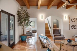 Uma sala de estar moderna com uma grande árvore em vaso, um sofá, uma poltrona, uma mesa de centro de vidro e uma escada que leva a outro andar.