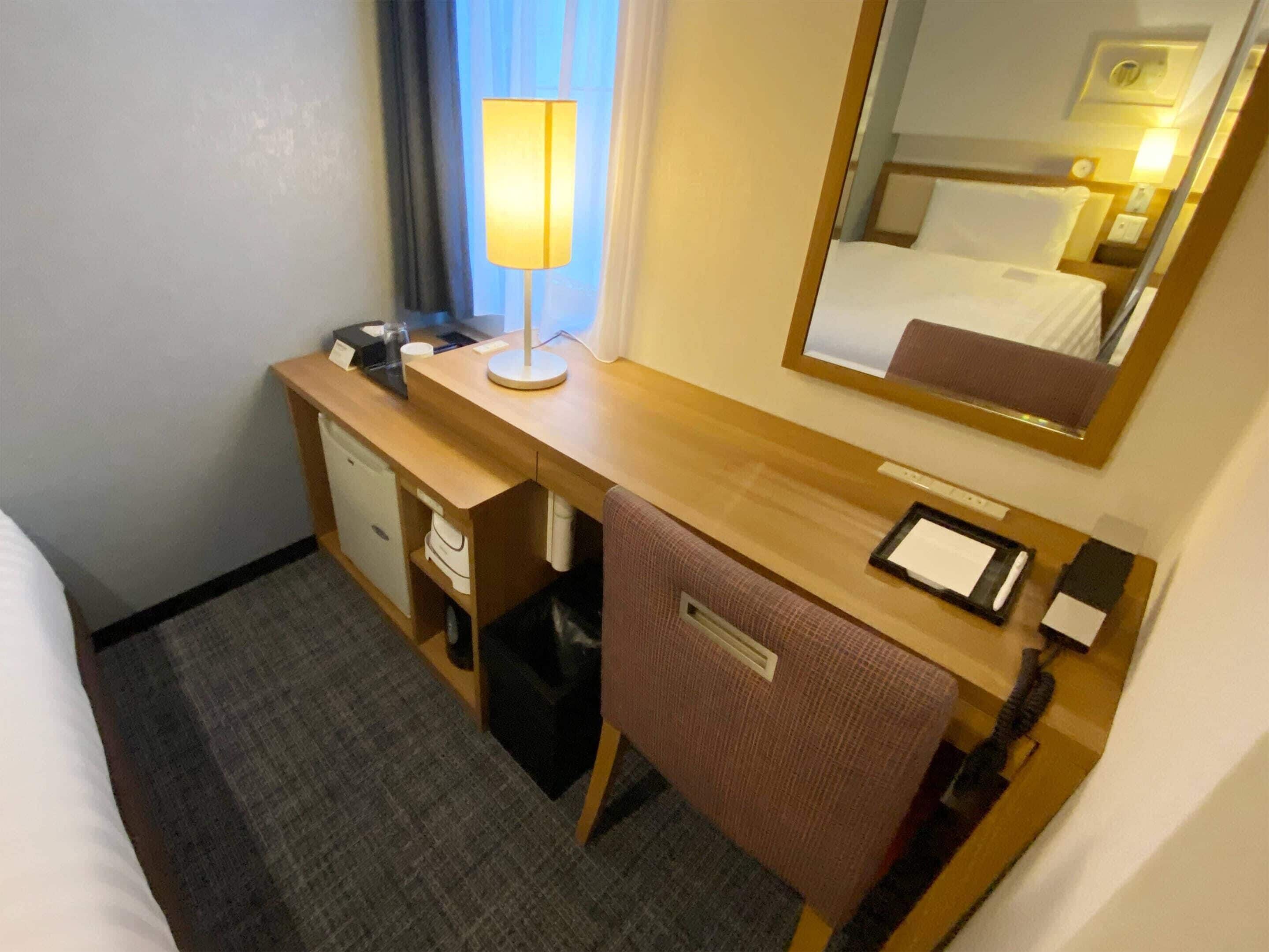 Habitación de hotel con un escritorio de madera, una silla, una cama, una mesita de noche, una lámpara, un espejo y un estante pequeño con un teléfono y un bloc de notas.