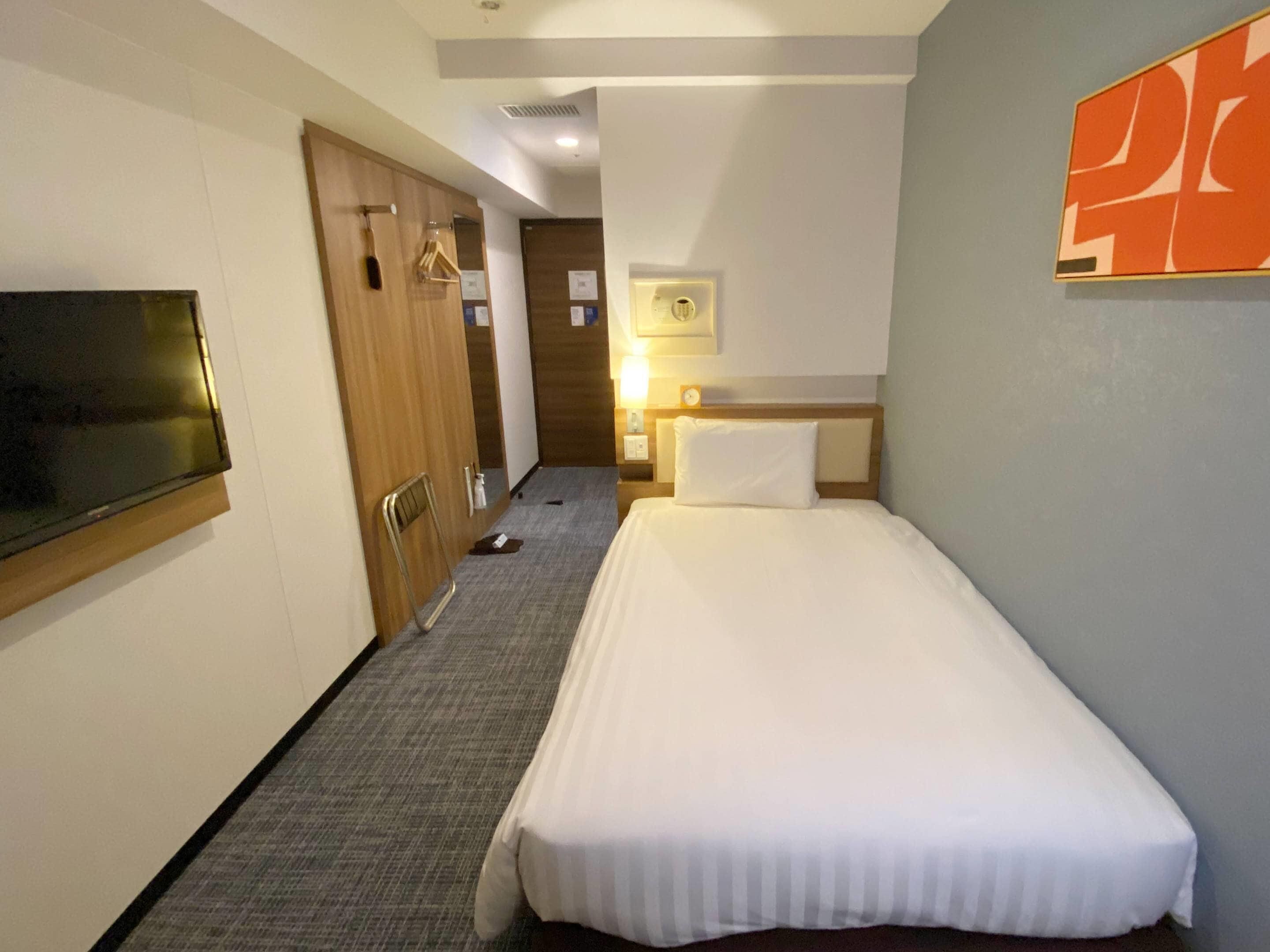 A hotel room with a single bed, a TV mounted on the wall, a wooden wardrobe, and a small desk with a lamp.
