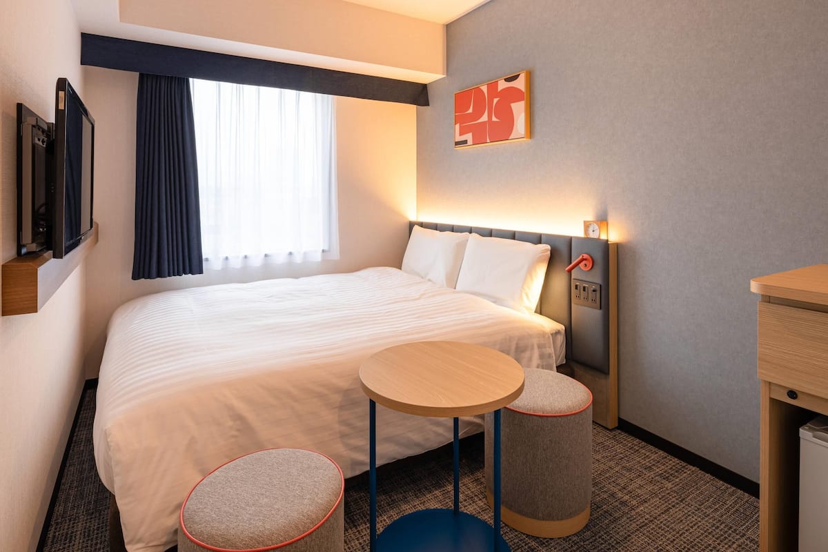 A hotel room with a large bed, a bedside table, two small stools, a TV, and a window with curtains.