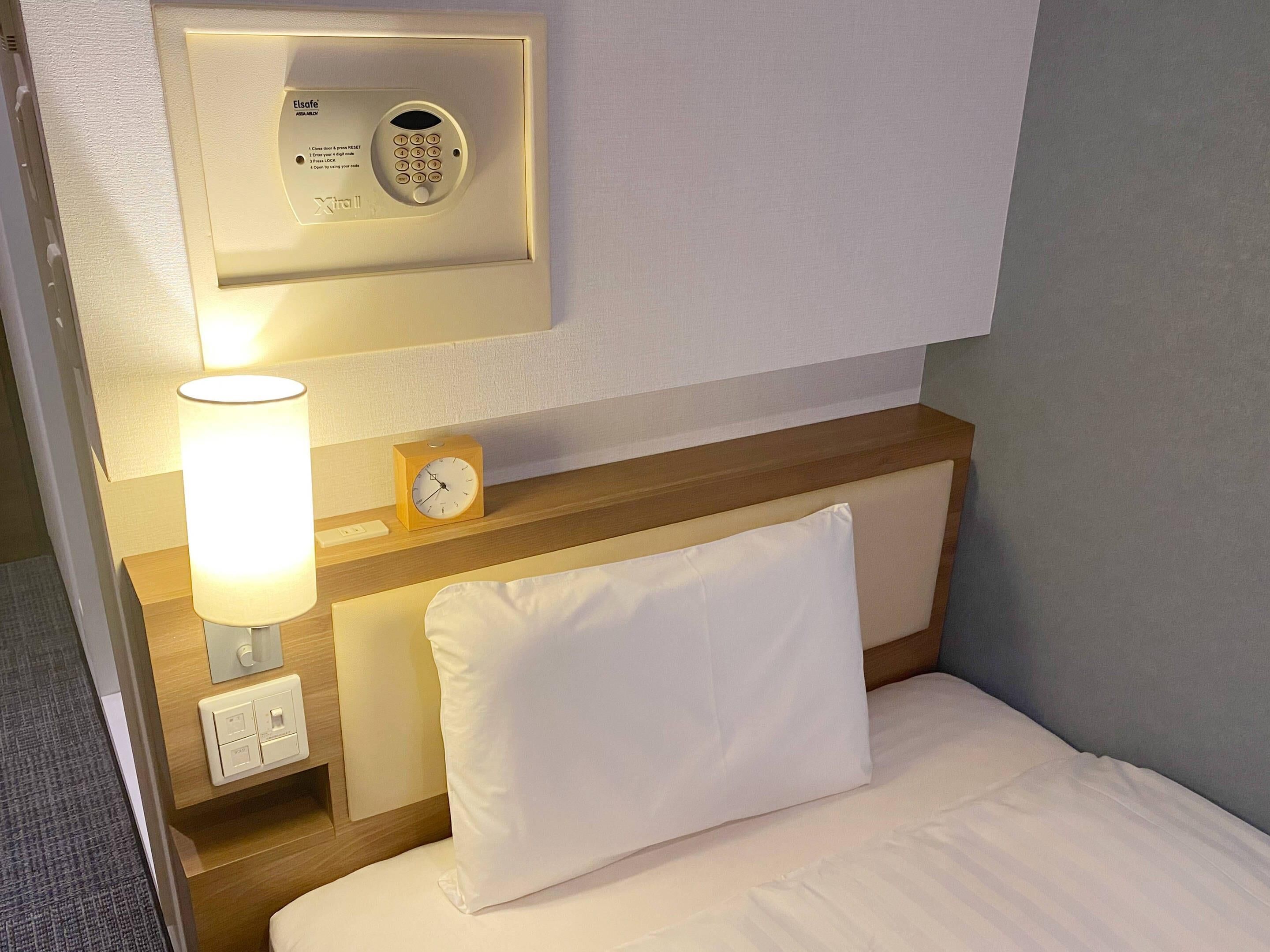 A hotel room with a bed, a nightstand, a lamp, a clock, and a wall-mounted control panel.