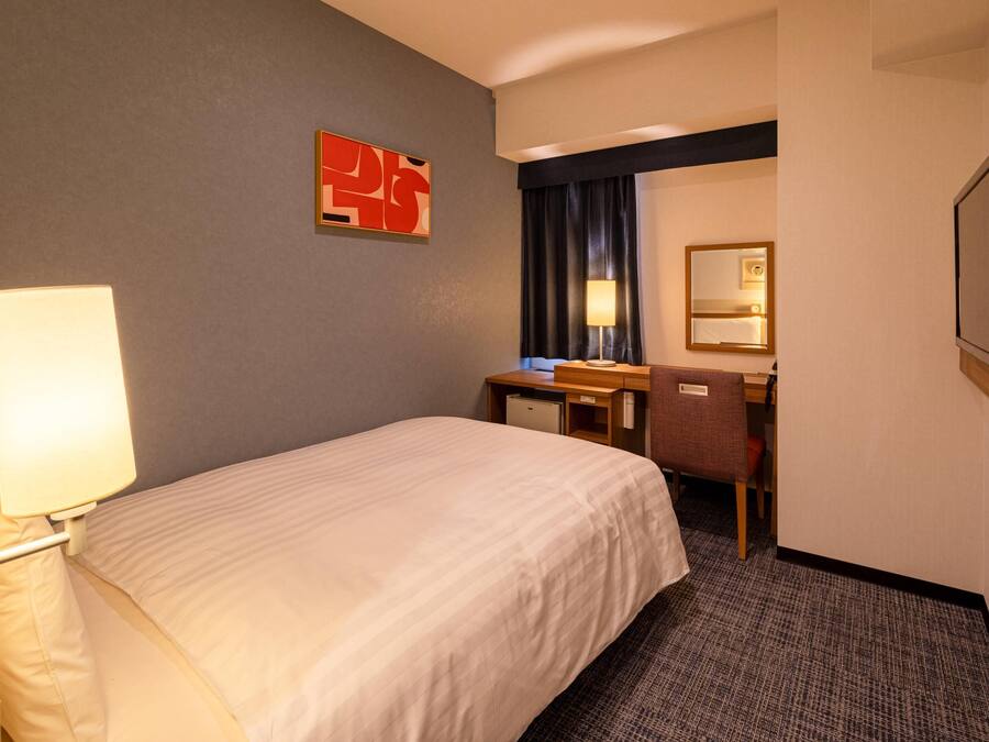 A hotel room with a bed, a desk, a chair, a lamp, a mirror, and a wall art piece.