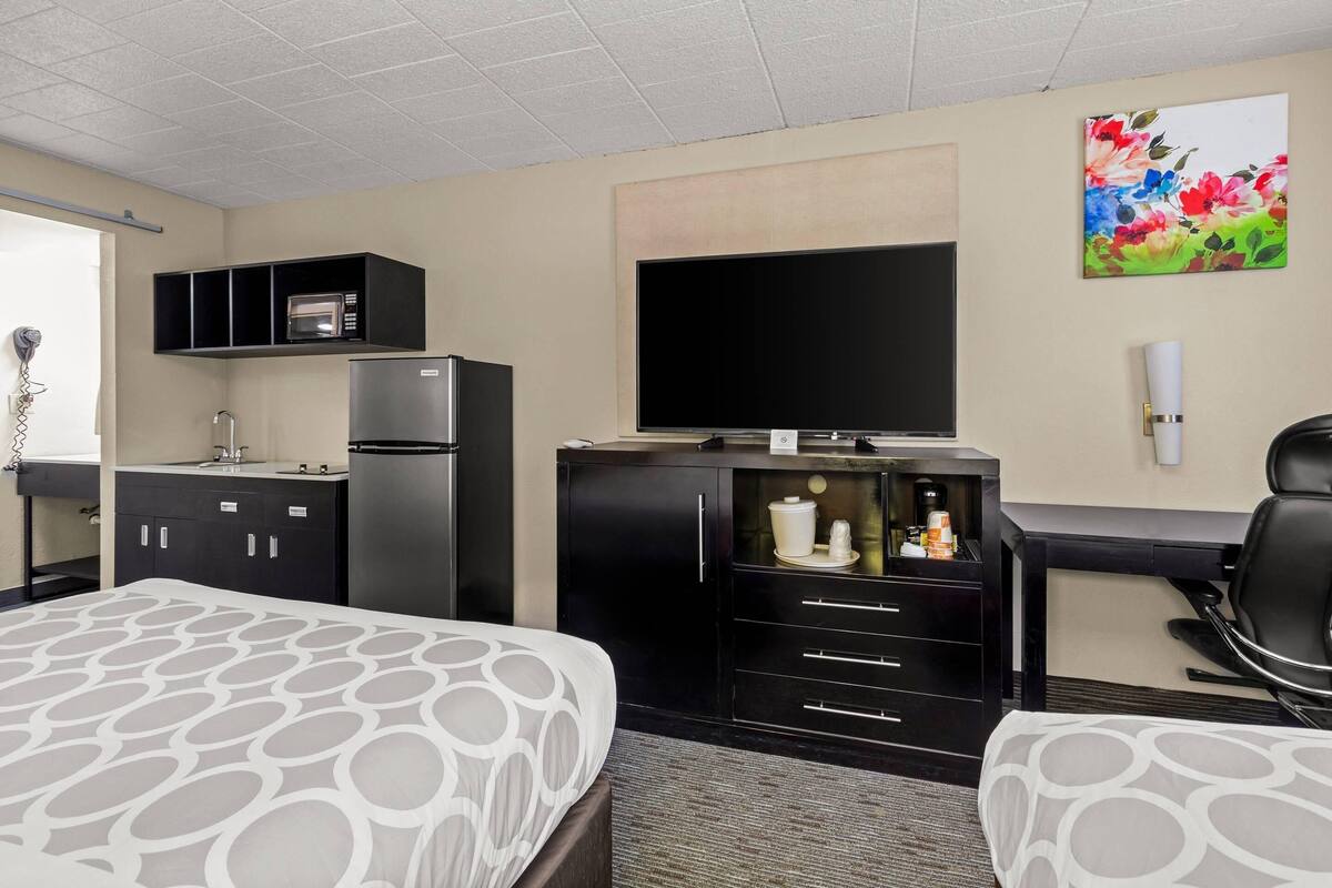 A hotel room with two beds, a kitchenette, a flat-screen TV, a painting, and a desk with a chair.