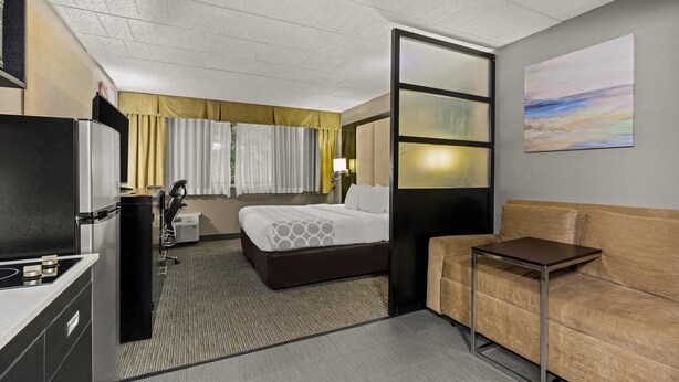 A hotel room with a bed, a sofa, a small table, a desk, a lamp, and a painting on the wall. - Suburban Studios Altoona (Altoona)