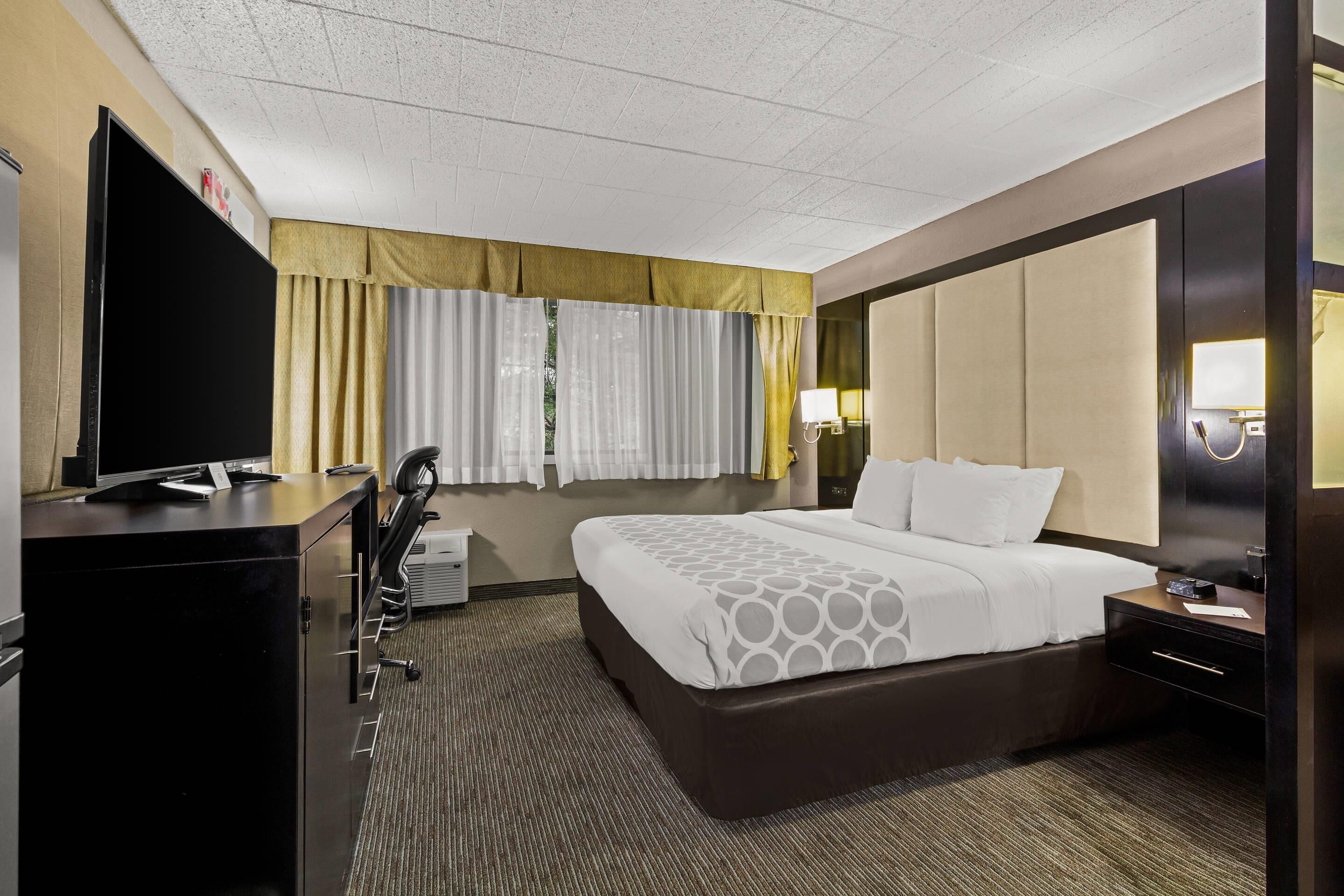 A hotel room with a large bed, a desk with a flat-screen TV, a chair, a desk lamp, and a nightstand.