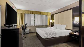 A hotel room with a large bed, a desk with a flat-screen TV, a chair, a desk lamp, and a nightstand.